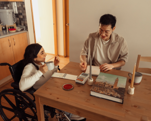 Una persona en silla de ruedas y una en silla sin ruedas sentadas en el comedor de su casa