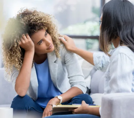 dos mujeres están sentadas hablando, una toca el hombro de la otra en señal de apoyo